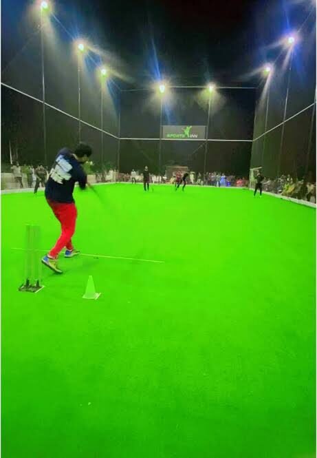 A batter plays a shot in a floodlit caged box-cricket ground at night